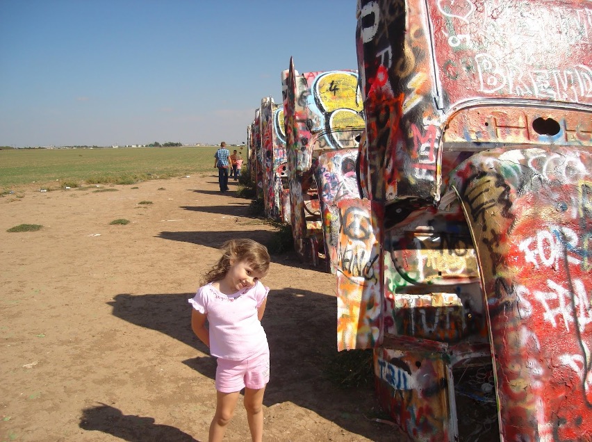 The author as a little girl in a light pink outfit posing happily in front of the Cadillacs.